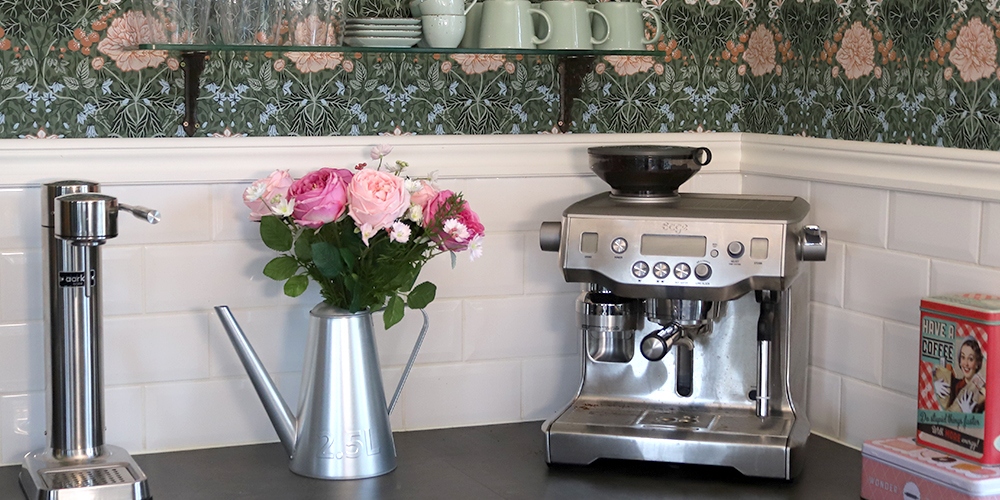 Ett stilrent kök med mörkgrön tapet med blommigt mönster och vitt kakel. På köksbänken står en bukett med rosa rosor i en silverfärgad vattenkanna, en sodastreamer i stål och en espressomaskin. En hylla ovanför rymmer kaffemuggar.