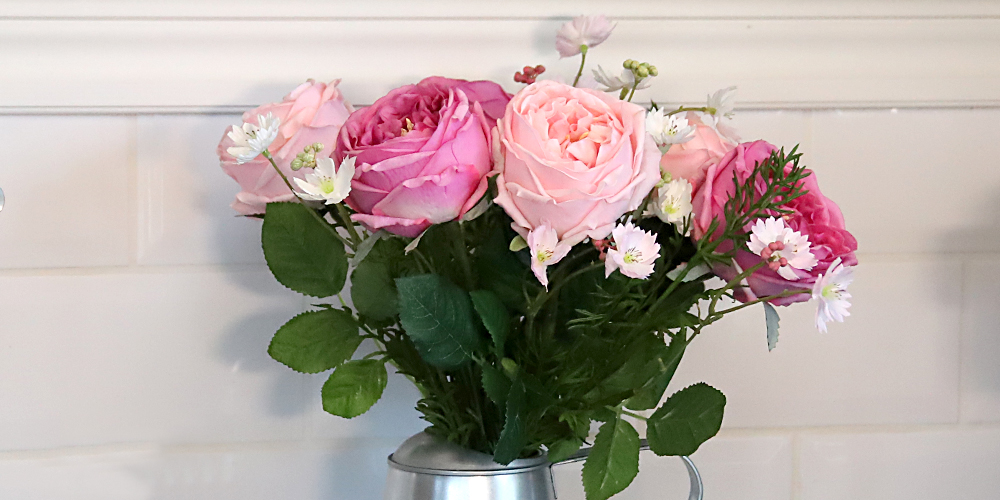 En närbild på en bukett med konstgjorda blommor i rosa och lila nyanser, tillsammans med små vita blommor och gröna blad. Blommorna står i en silverfärgad vas mot en bakgrund av vitt kakel och en vit vägglist.