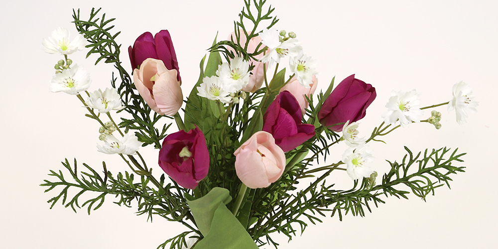 En bukett med konstgjorda blommor, bestående av lila och ljusrosa tulpaner, små vita blommor och frodiga gröna barrliknande kvistar, mot en ljus bakgrund.