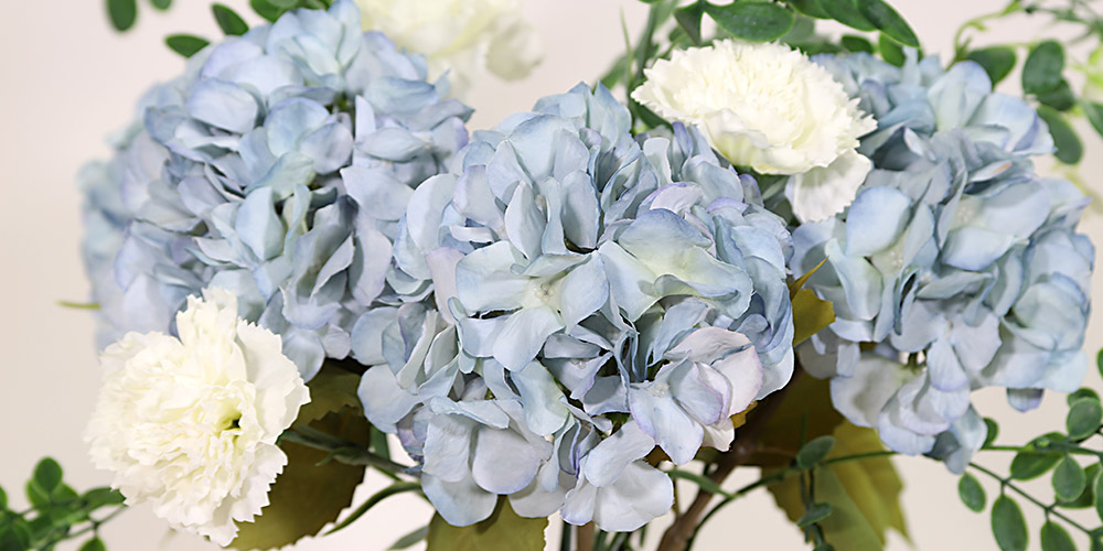 En närbild på en bukett med konstgjorda ljusblå hortensior med fylliga blomhuvuden, blandade med vita nejlika blommor och gröna blad.